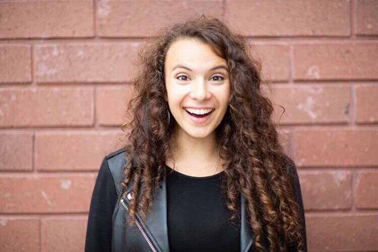 This is an image of Lyndsay smiling wide in front of a brick wall wearing a black leather jacket.